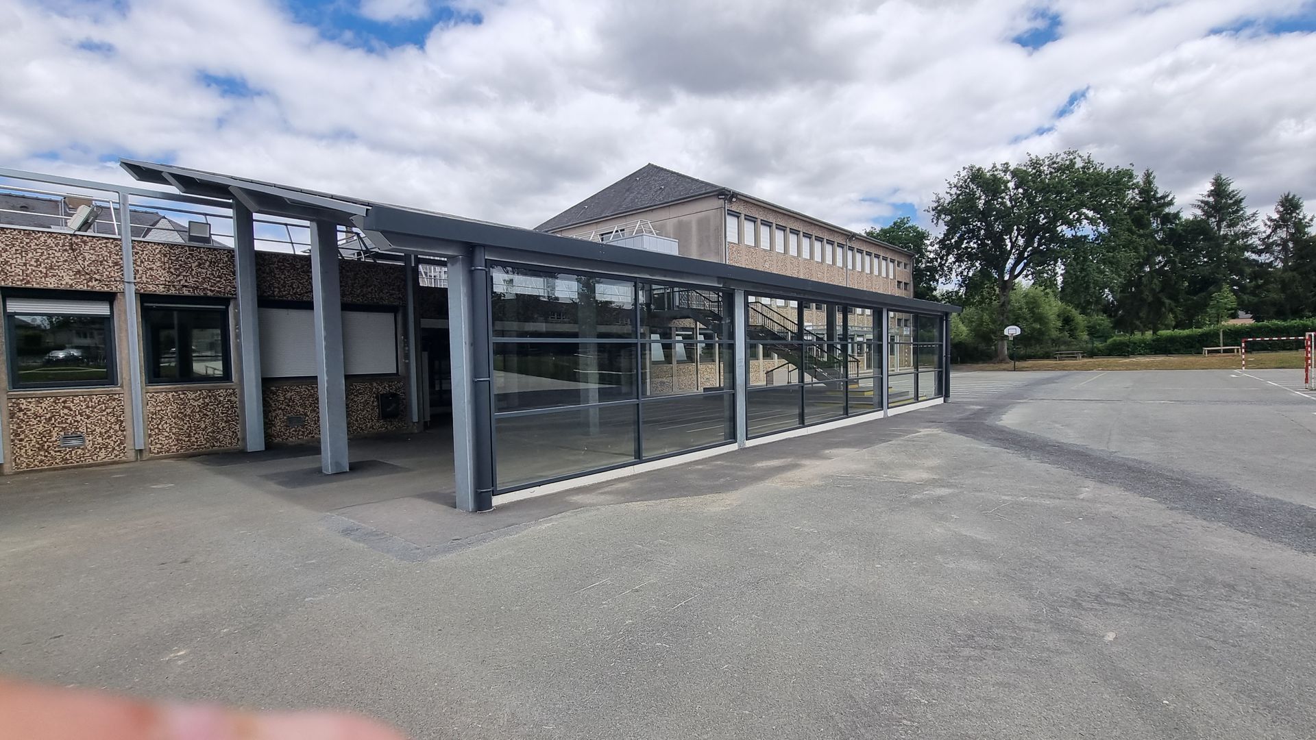 Une allée couverte aux parois de verre, attenante à un bâtiment beige, sur une cour d'école pavée, sous un ciel partiellement nuageux.