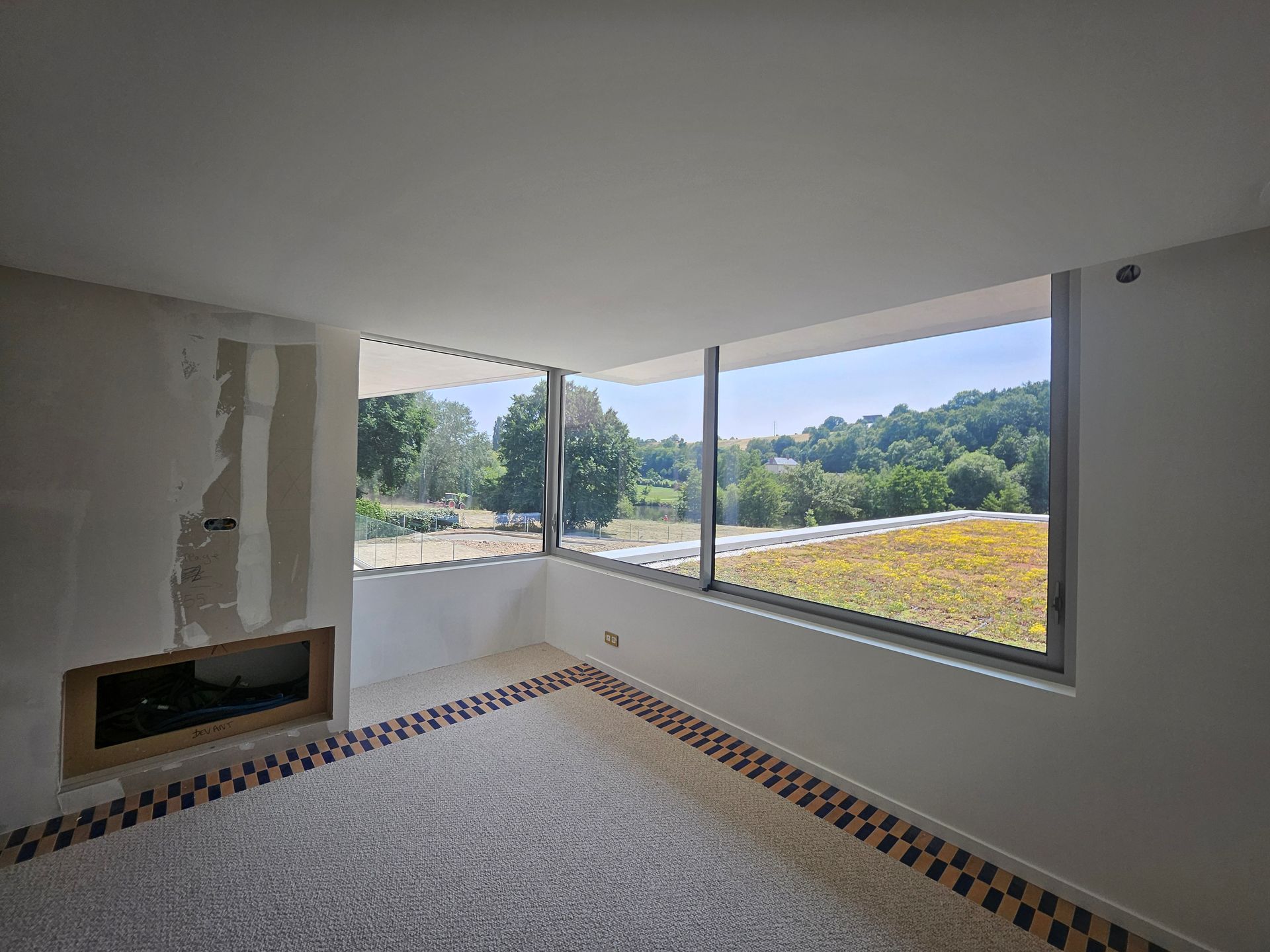 Une pièce inachevée dotée d'une cheminée minimaliste, d'une bordure de sol texturée et de grandes fenêtres donnant sur un paysage verdoyant.