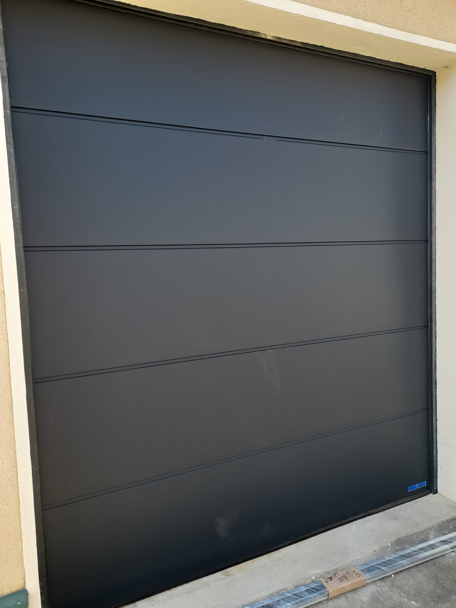 Porte de garage sectionnelle moderne gris foncé, composée de quatre panneaux horizontaux encadrés d'un stuc clair.