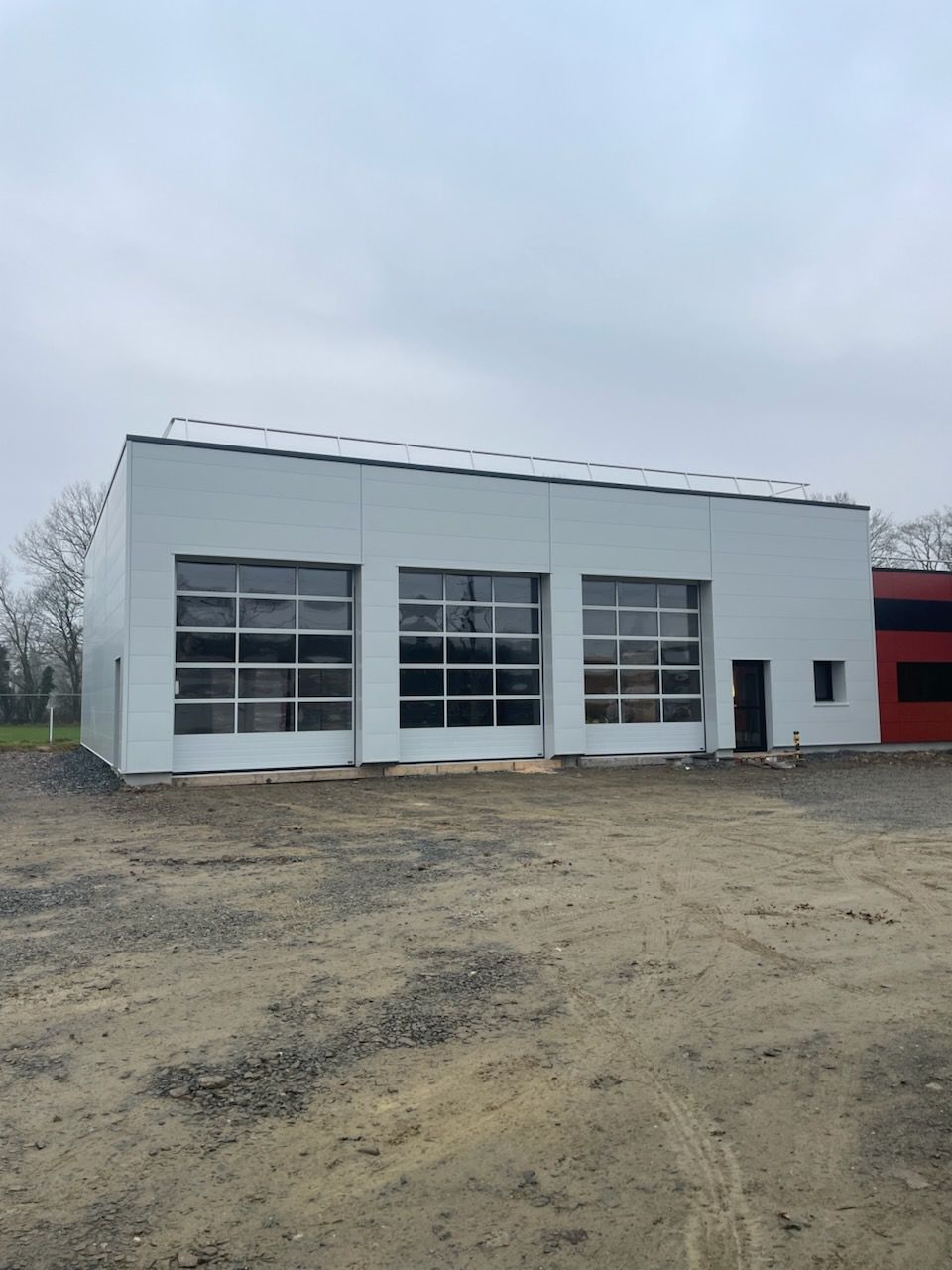 Un bâtiment industriel blanc moderne avec trois grandes portes de garage vitrées, une seule porte d'entrée et un terrain non pavé.