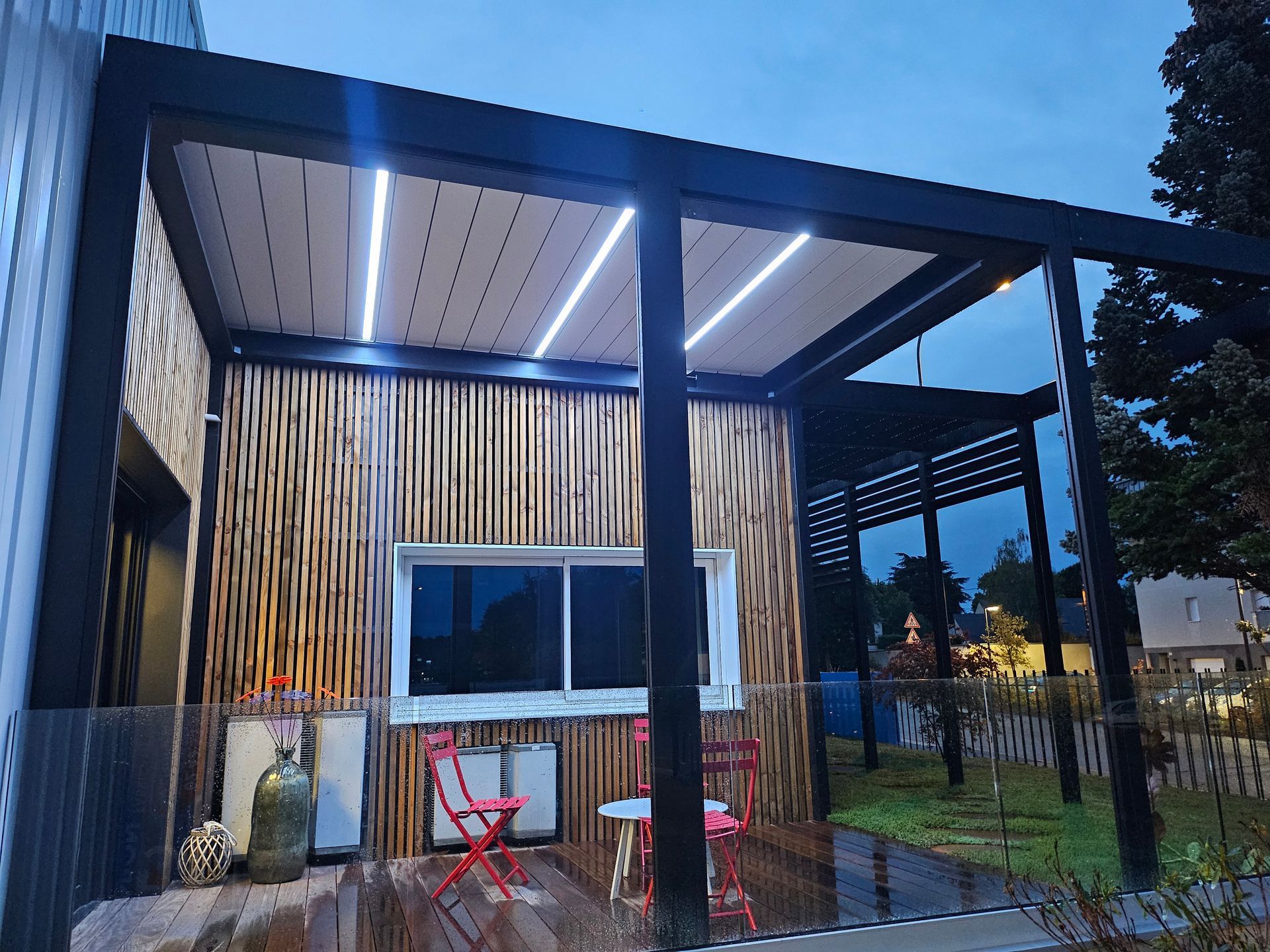 Terrasse moderne avec toit rétractable intégrant un éclairage LED, un bardage en bambou et des chaises de bistrot rouges au crépuscule.
