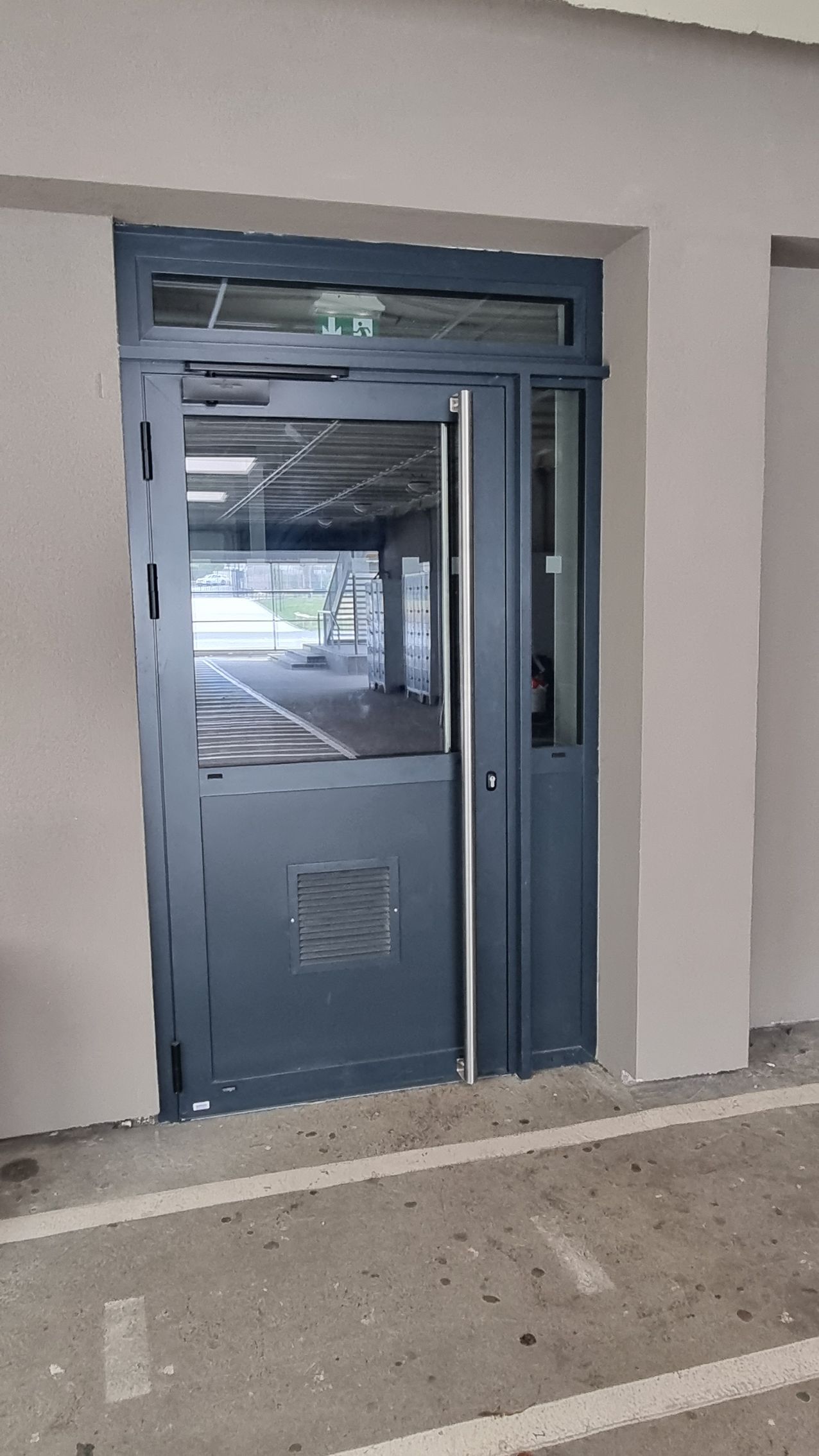 Une porte en métal gris avec une vitre et une grille d'aération inférieure, intégrée à un mur beige, donne accès à un espace couvert.