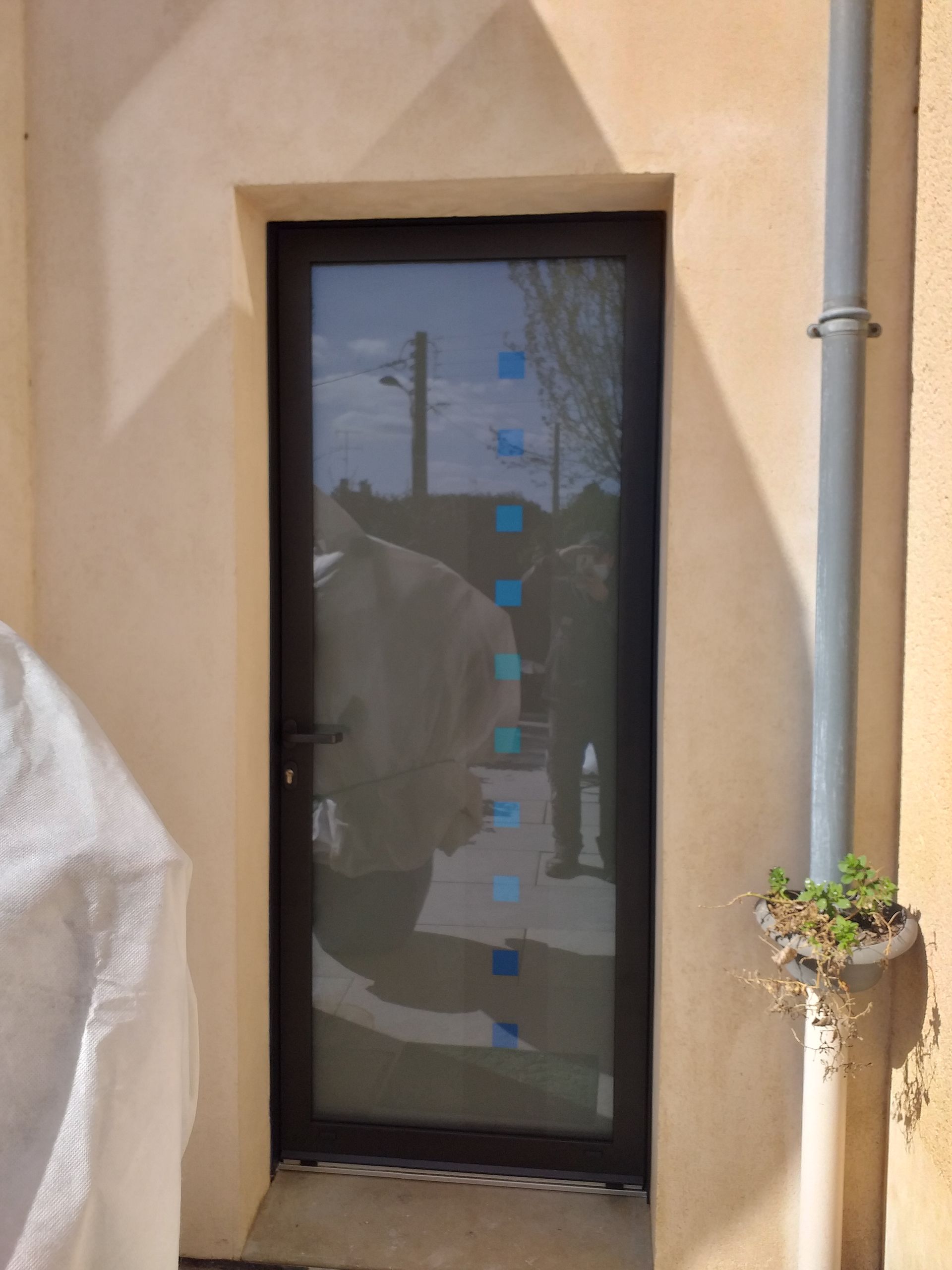 Une haute porte vitrée à cadre sombre, encastrée dans un mur en stuc clair, ornée d'une colonne verticale de petits autocollants carrés bleus.