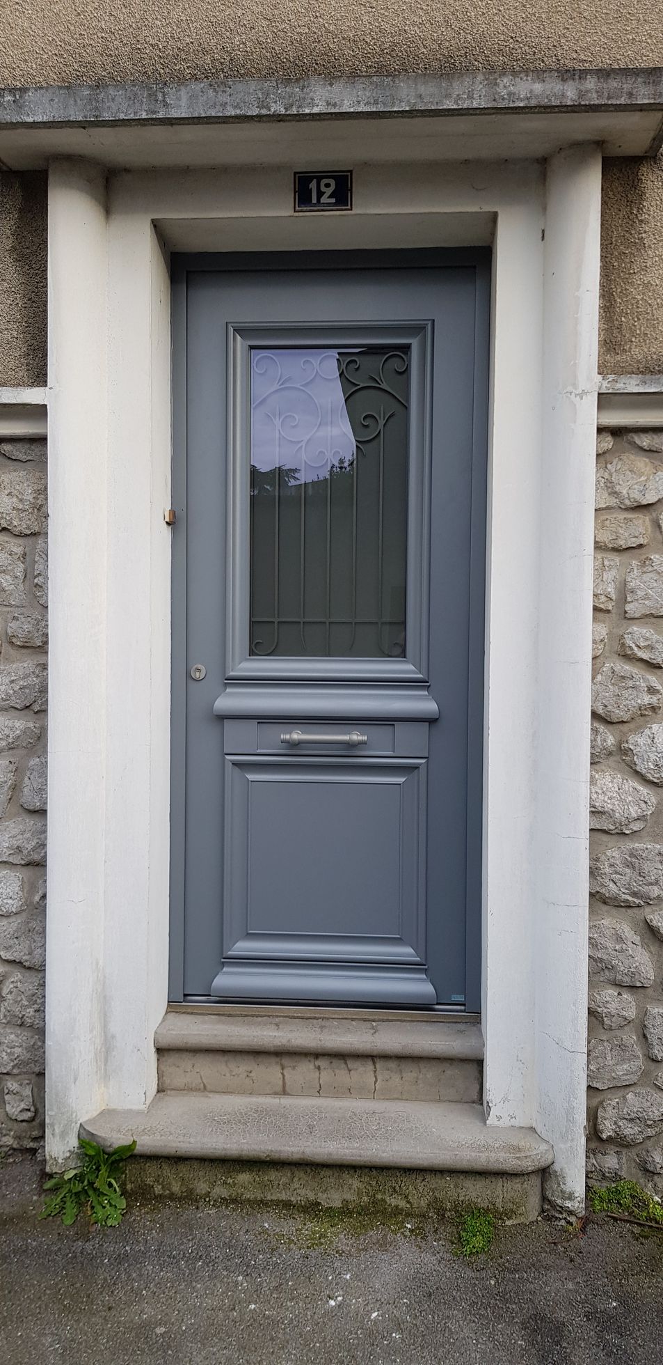 Une porte d'entrée grise avec un seul panneau de verre et le numéro 12 au-dessus, encastrée dans un mur de pierre avec des encadrements blancs et des marches en pierre.