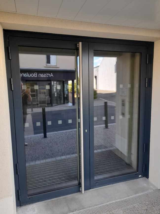 Portes d'entrée à double vitrage dans un cadre en métal gris foncé, reflétant une scène de rue extérieure avec une boulangerie artisanale.
