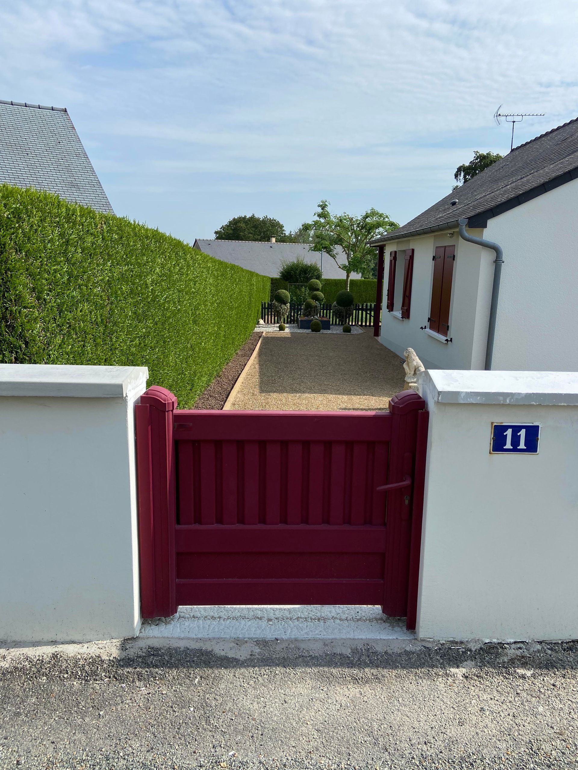 Un portail bordeaux entre deux piliers blancs, portant le numéro 11, menant à un chemin de gravier longeant une haie verte et une maison.