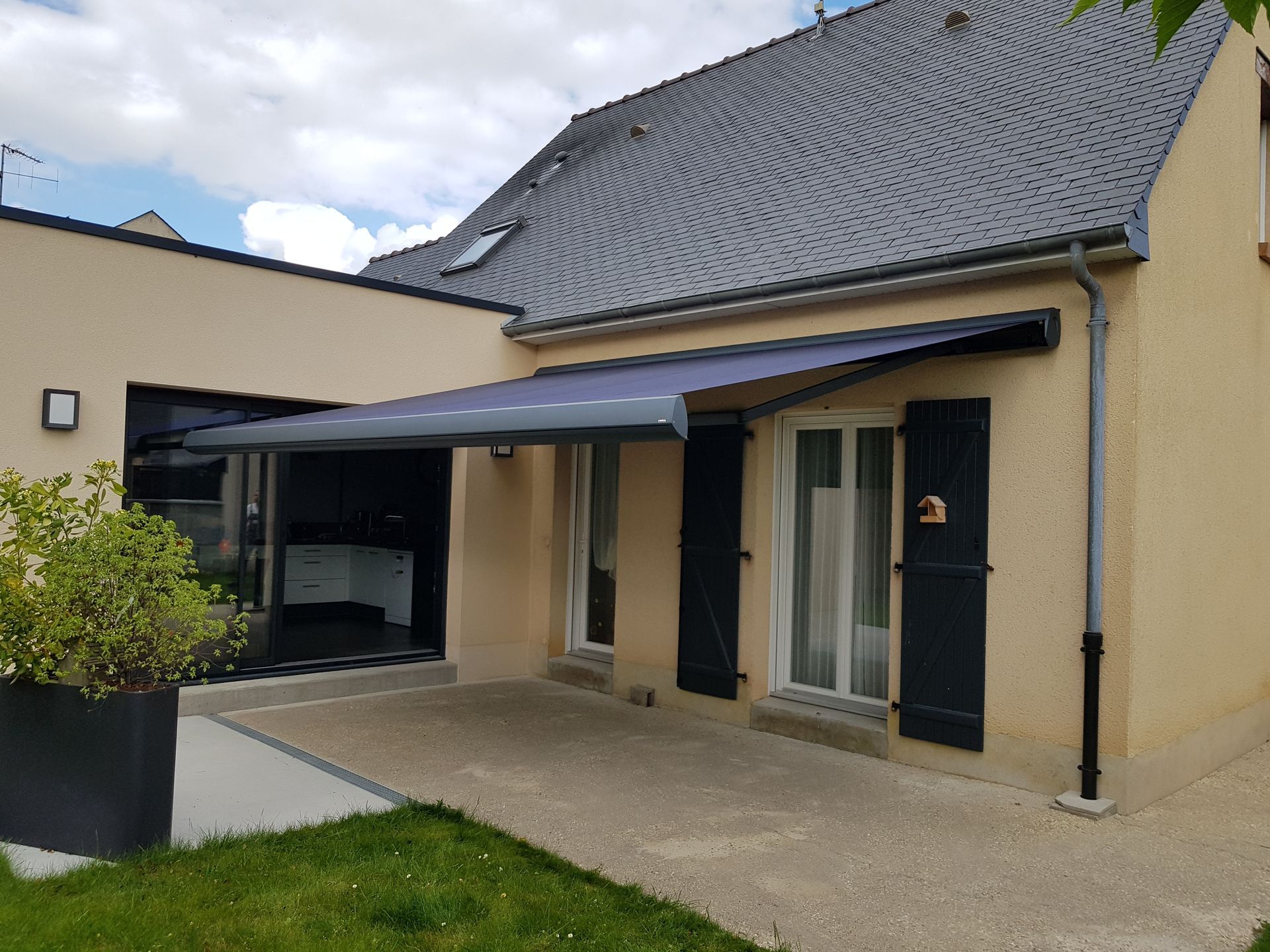 Une maison beige avec un auvent gris surplombant une terrasse en pierre et un pot de fleurs noir au premier plan.