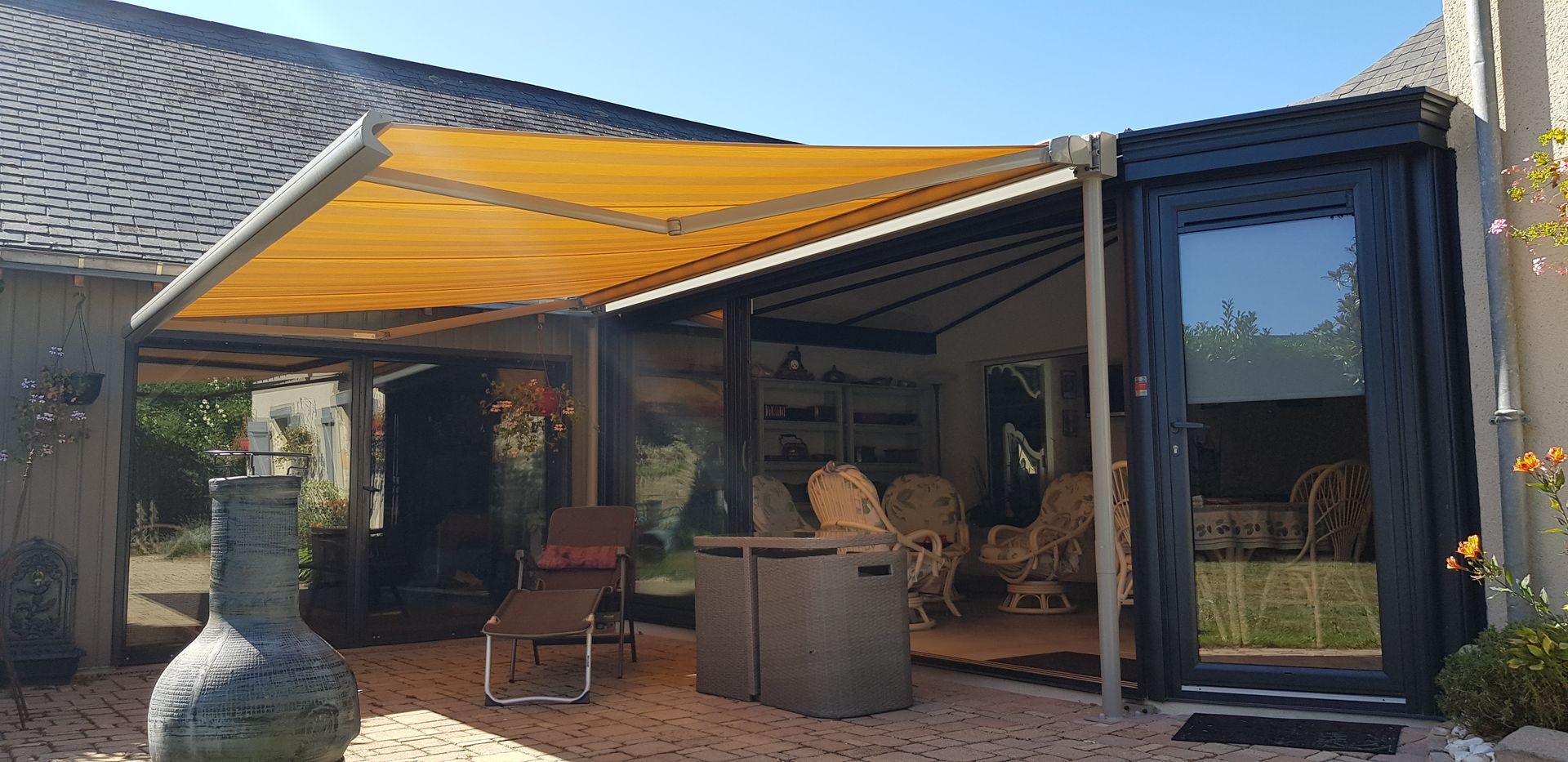 Un store rétractable jaune vif ombrage une terrasse meublée de chaises en osier et d'une chaise longue posée sur du gravier.