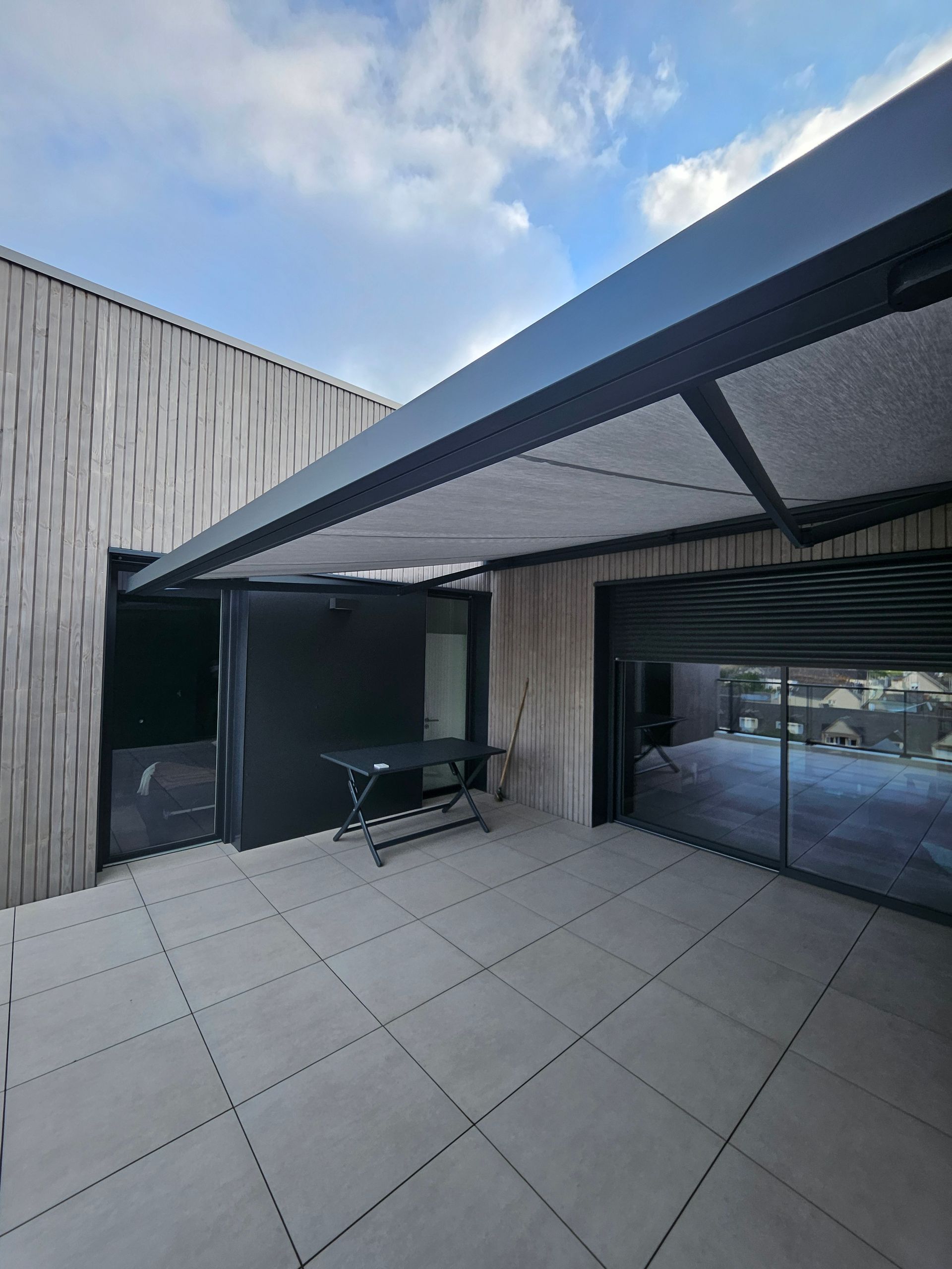 Une terrasse moderne sur le toit avec des carreaux gris clair, des murs extérieurs gris foncé, un mur en bois à claire-voie et un auvent rétractable.