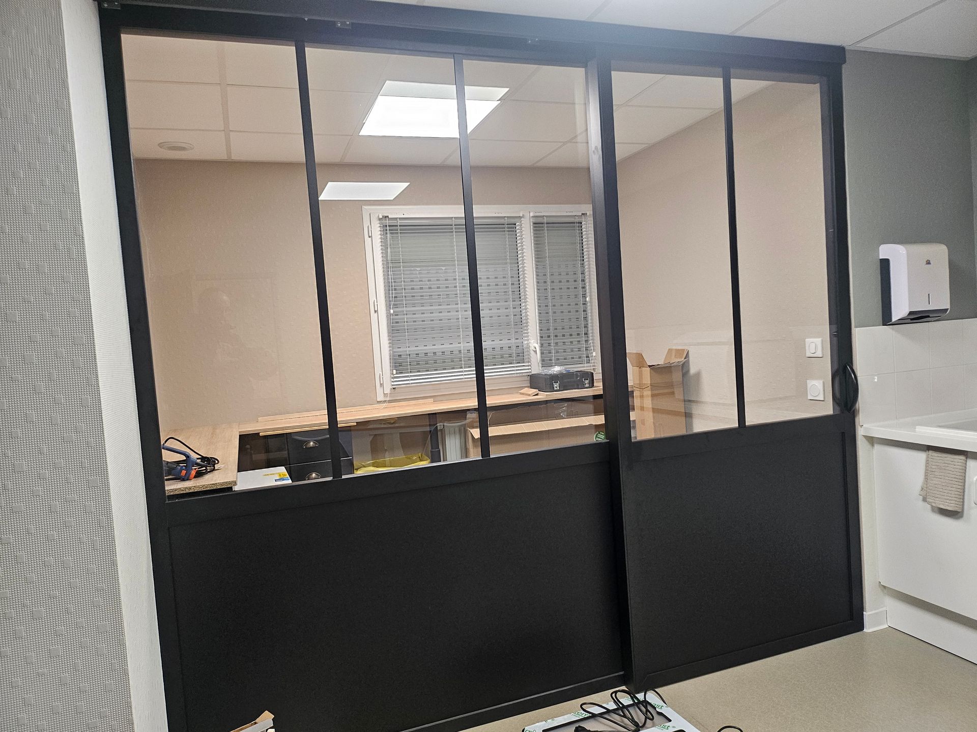 Une cloison coulissante en verre noir de style industriel sépare un espace de bureau avec un bureau et une fenêtre visibles à l'intérieur.