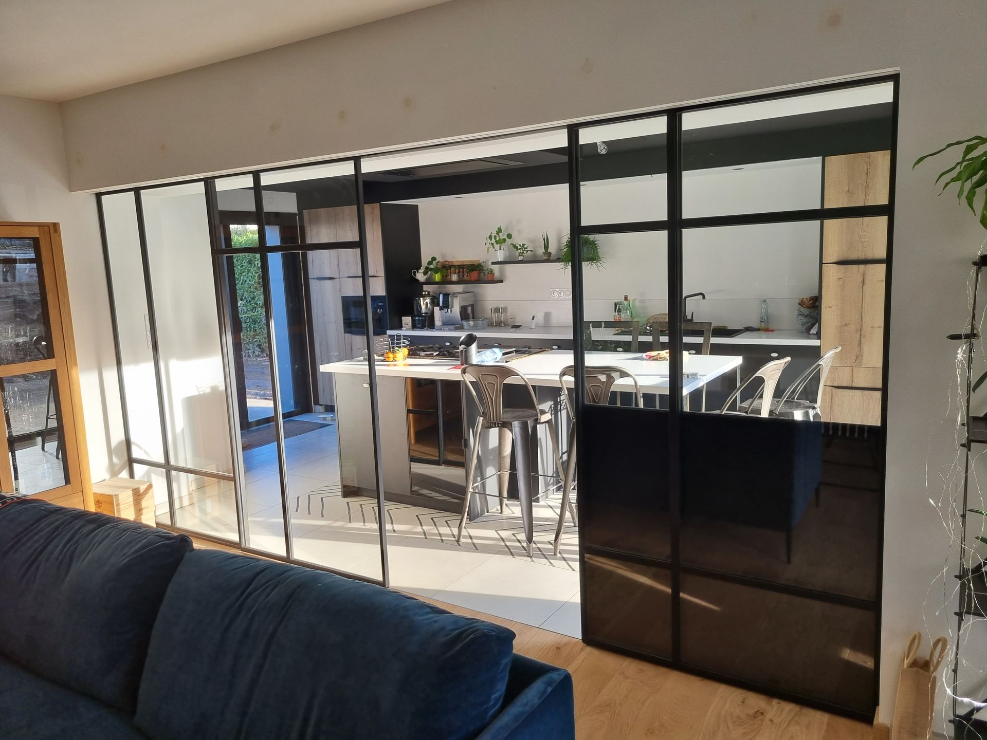 Vue du salon vers la cuisine, séparée par une cloison vitrée à cadre noir.