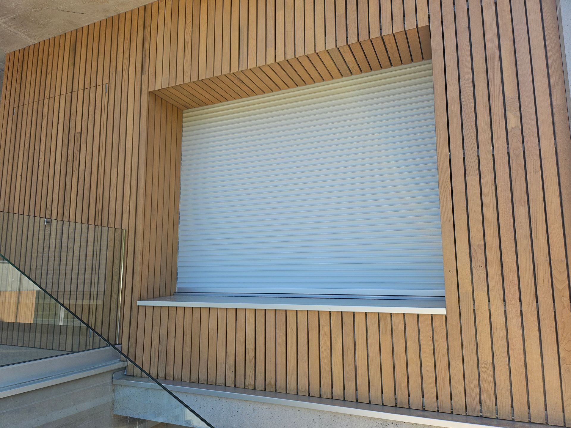 L'extérieur d'un bâtiment présente des lattes verticales en bois clair et un volet roulant blanc fermé.