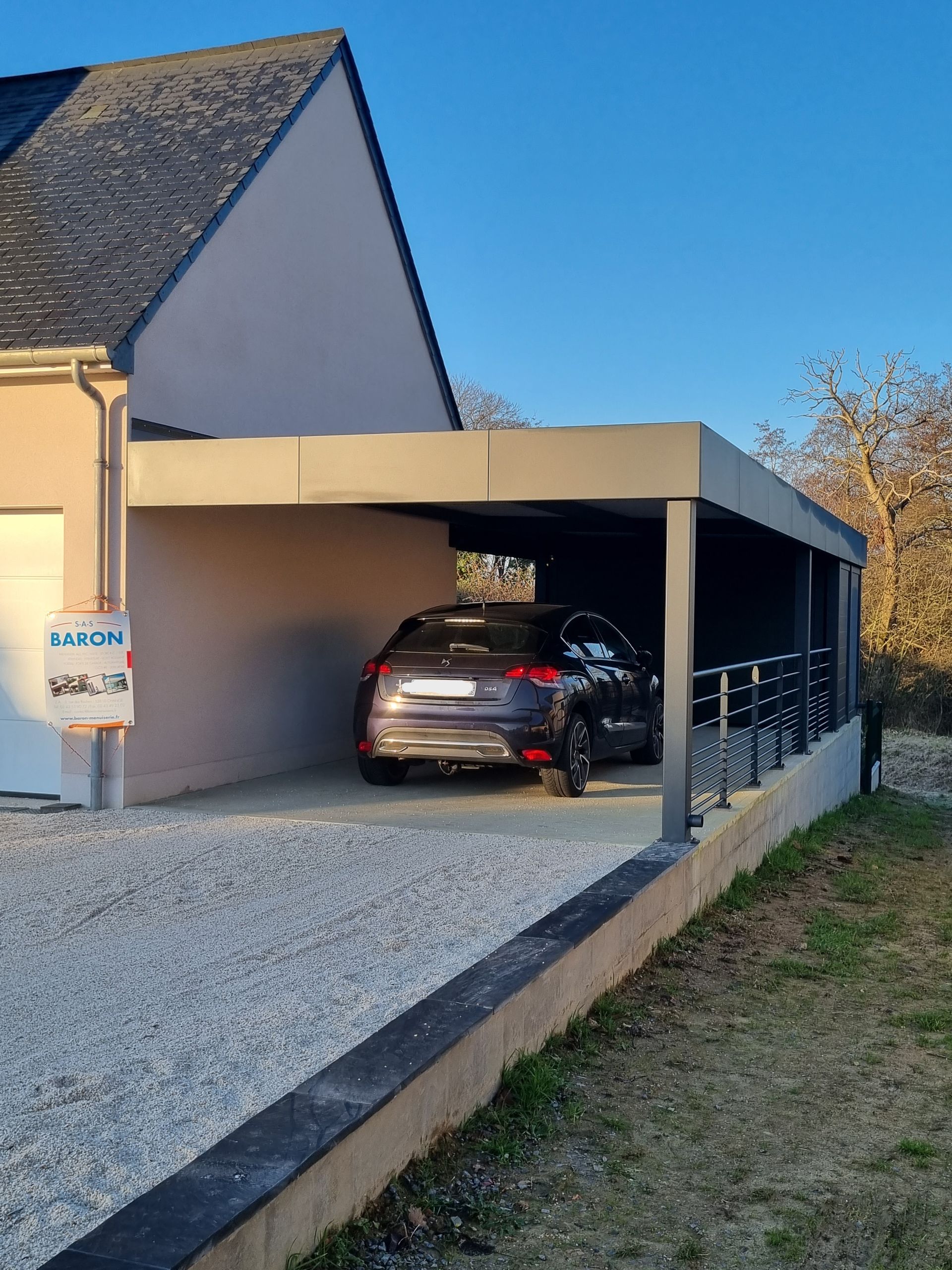 Une voiture sombre est garée sous un abri de voiture moderne en métal à toit plat, adossé à une maison de couleur claire.