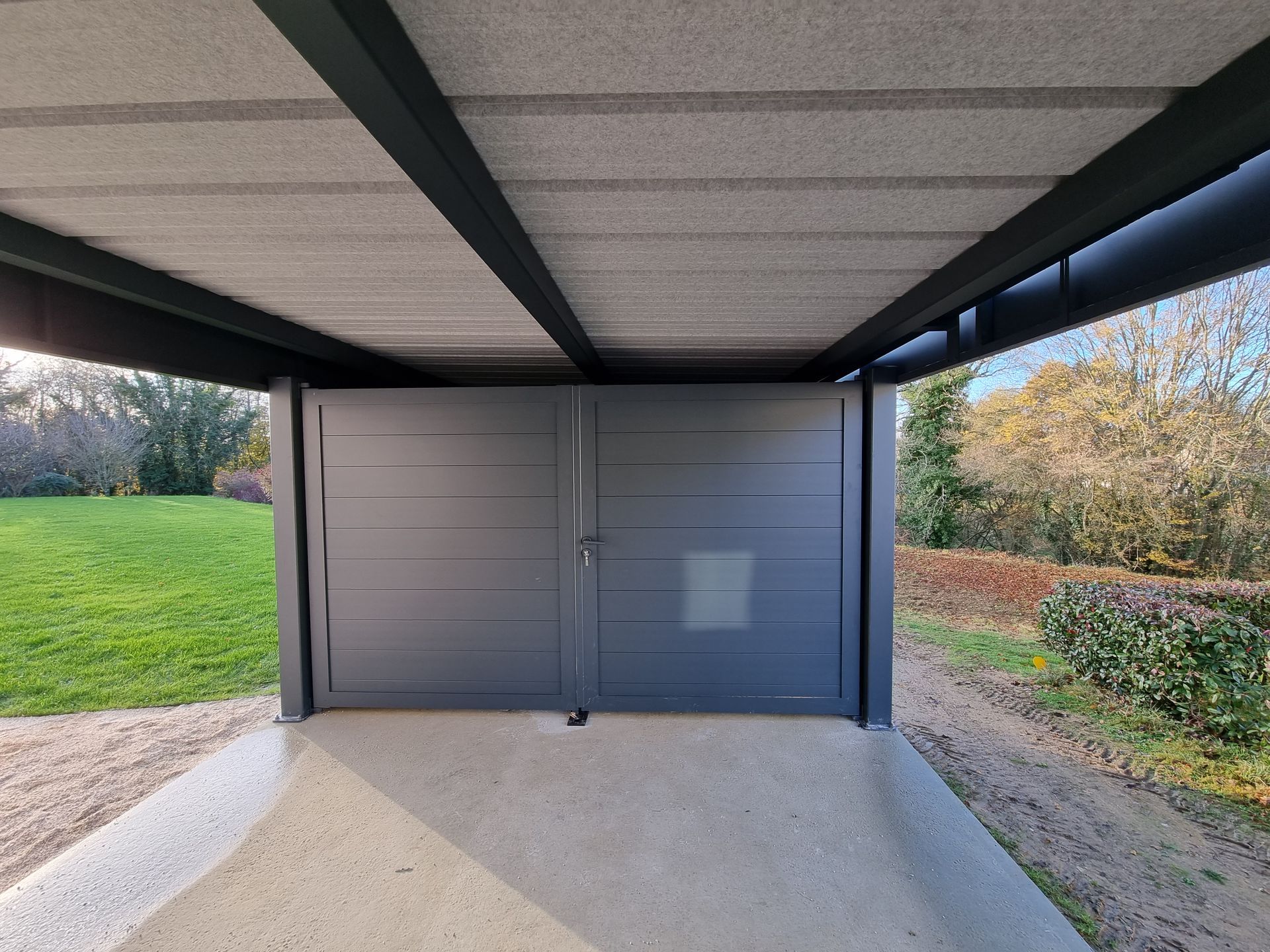 Un portail d'allée moderne gris à double battant, installé sous un abri de voiture couvert, adossé à une pelouse et à des arbres.