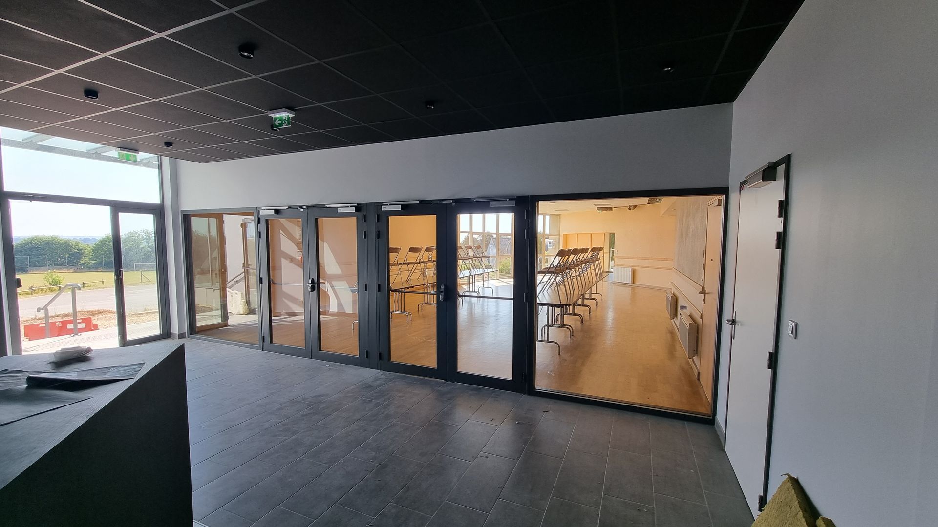Une pièce moderne avec un sol carrelé gris, des murs blancs, un plafond en dalles noires et de grandes cloisons vitrées donnant sur une salle de sport.