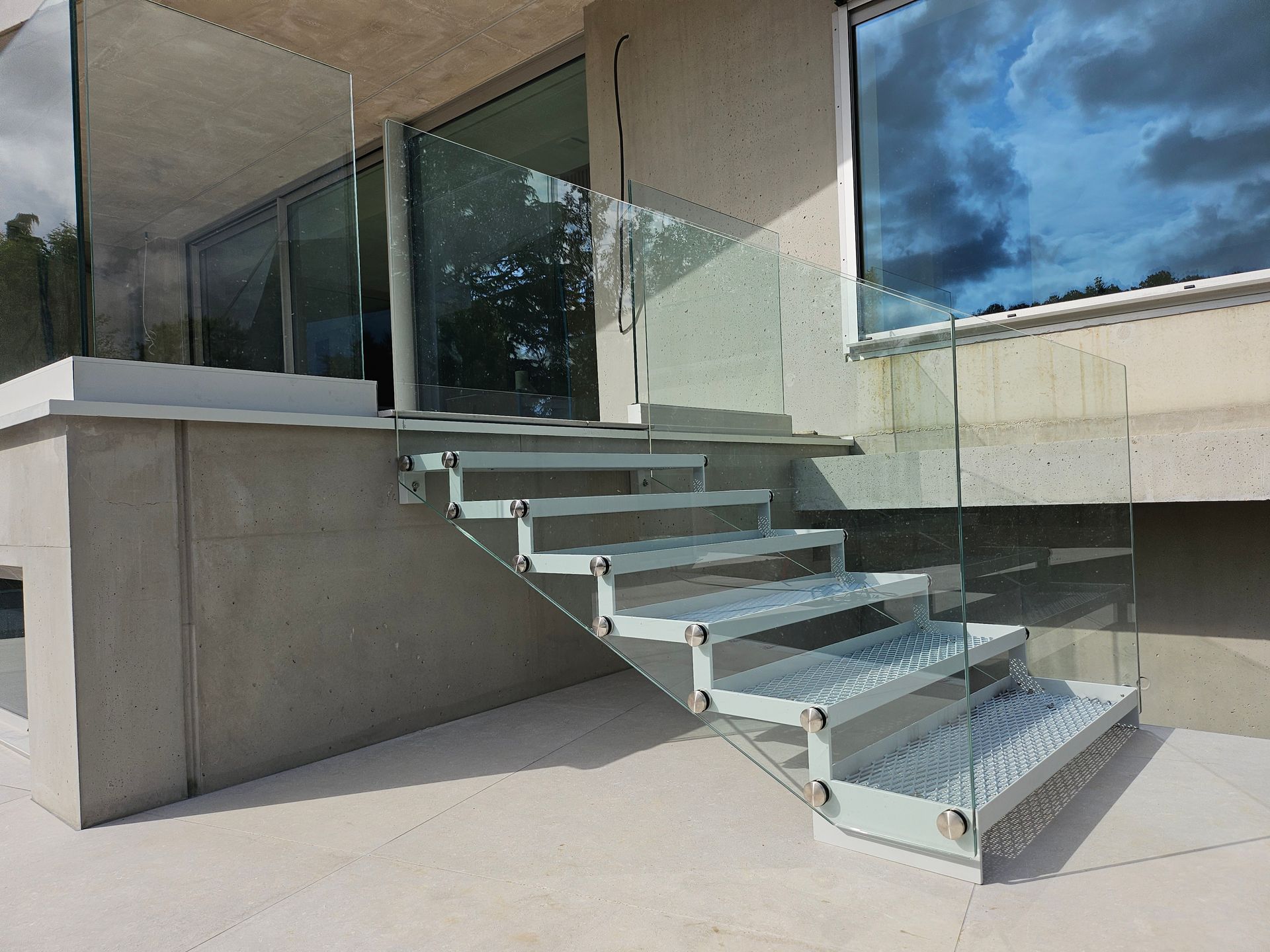 Un escalier extérieur moderne avec des garde-corps latéraux en verre et des marches gris clair, menant à une maison avec un mur en béton.