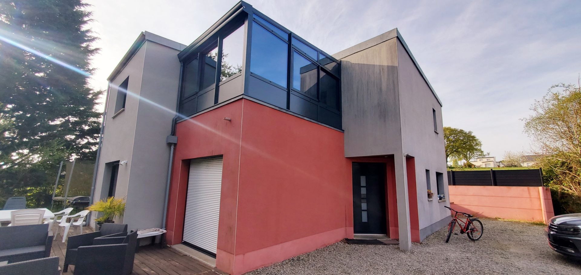 Une maison moderne de deux étages avec un enduit gris et rouge, de grandes fenêtres à l'étage, une porte sectionnelle blanche et un patio.
