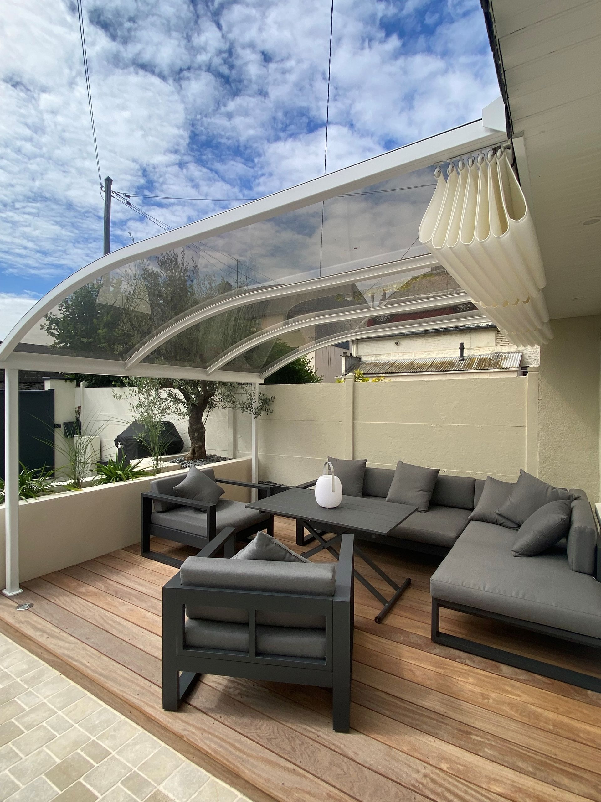 Une terrasse extérieure moderne avec des meubles de salon gris sur une terrasse en bois sous un auvent incurvé partiellement rétracté.