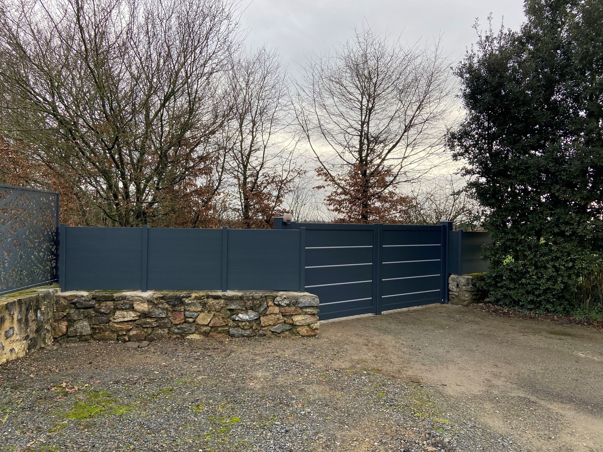 Un portail d'allée en métal bleu foncé et des panneaux de clôture assortis se trouvent derrière un muret en pierre rustique, dans une zone boisée.