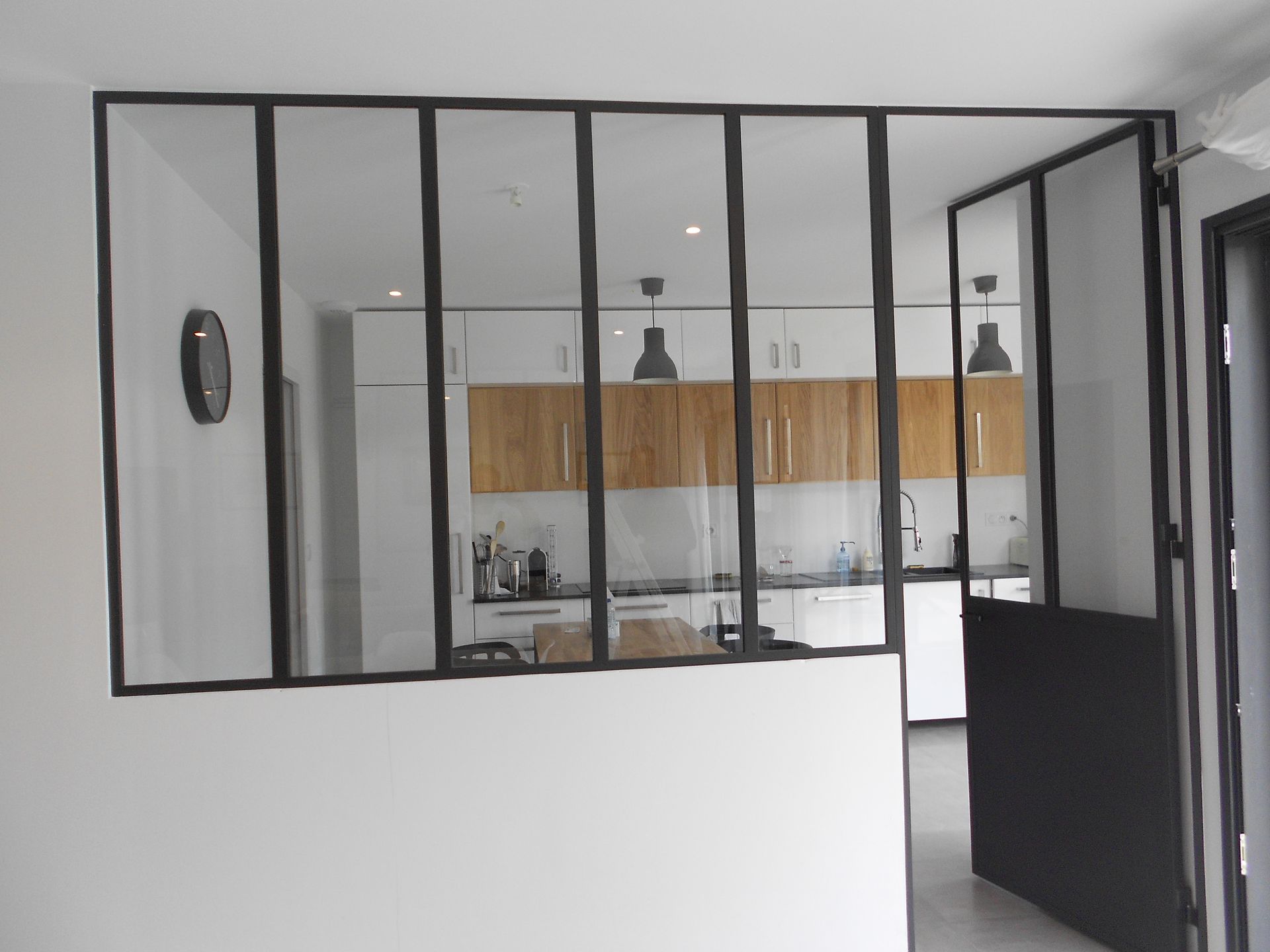 Une cloison en métal noir et en verre de style industriel sépare un espace de vie d'une cuisine moderne avec des placards en bois.