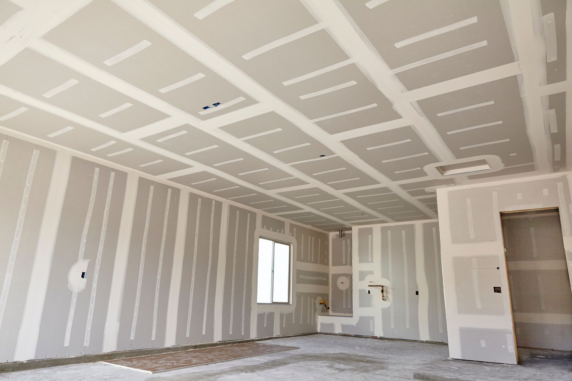Mann mit gelbem Schutzhelm trägt Trockenbauspachtelmasse auf eine Wand in einem unfertigen Raum auf.