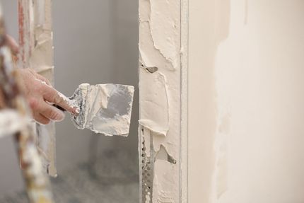 Eine Person mit einem orangefarbenen Handschuh glättet mit einer Kelle nassen Zement an einer Wand.