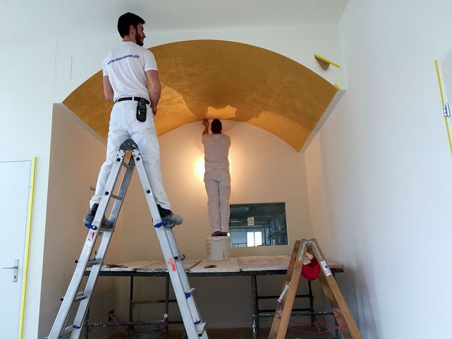 Zwei Maler tragen Blattgold auf eine gewölbte Decke auf. Einer auf einer Leiter, der andere auf einem Eimer in einem Raum.