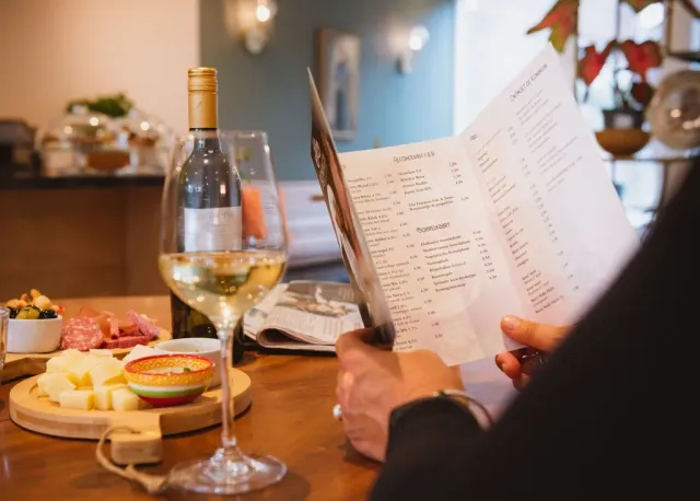 Iemand die een menukaart leest aan een tafel gedekt met wijn, kaas en vleeswaren.
