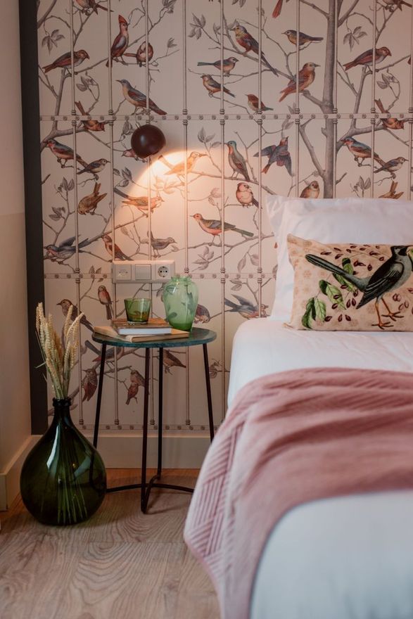 Slaapkamer met behang met vogelmotief, een klein bijzettafeltje, een bed met een roze sprei, een groene vaas en een lamp.