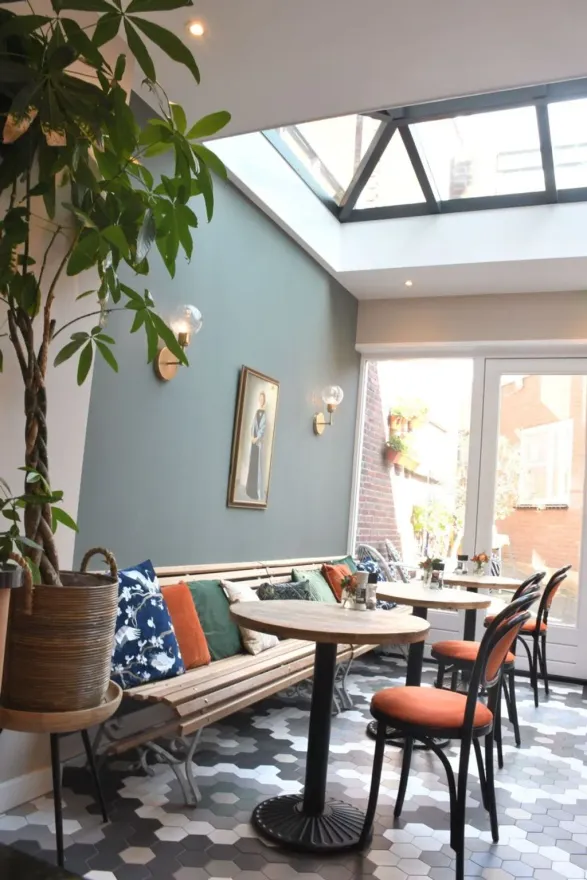 Café-interieur met groene muur, dakraam, zitbanken met kussens en ronde tafels.