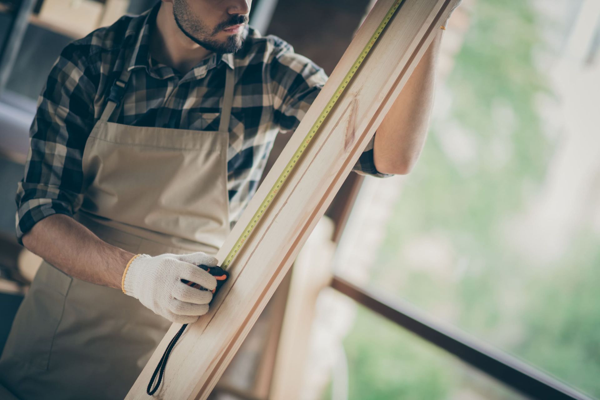 Carpintero con guantes y delantal midiendo una tabla de madera con una cinta métrica.