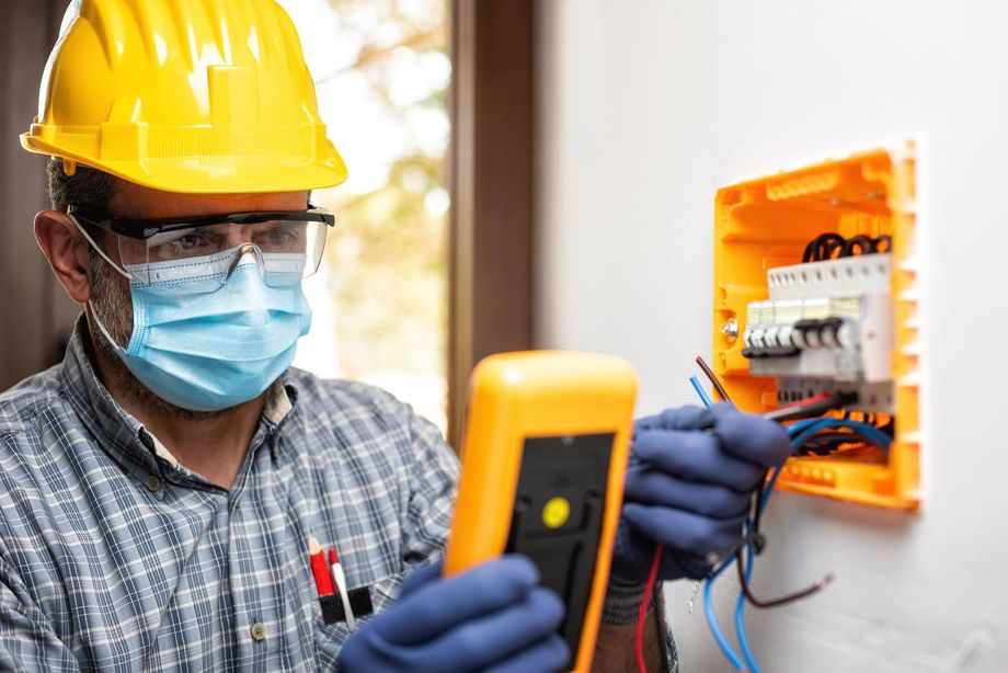 Electricista con casco, máscara y guantes, probando el panel eléctrico con multímetro.