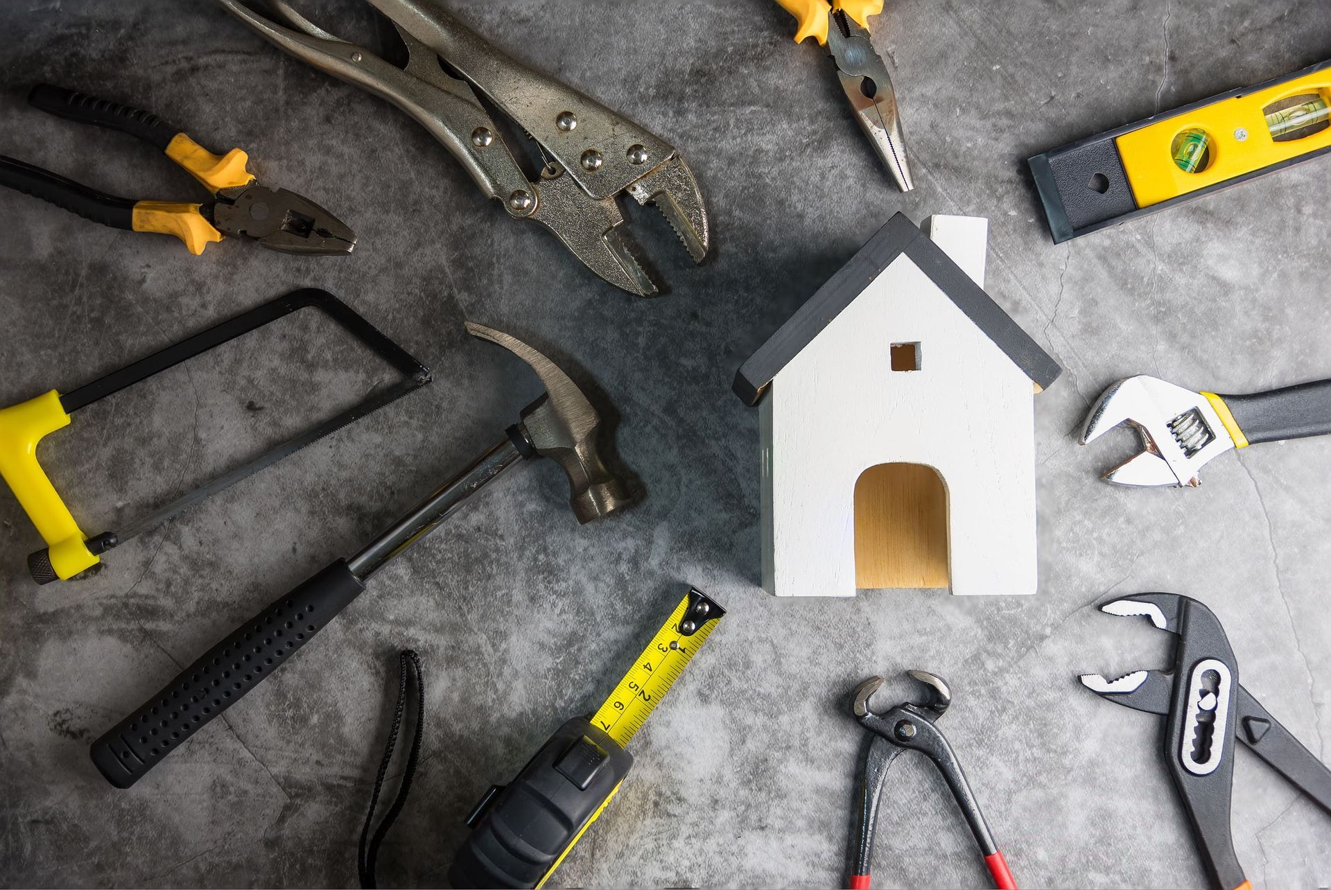 Una casa modelo rodeada de varias herramientas sobre una superficie gris, que representa la reparación y construcción del hogar.