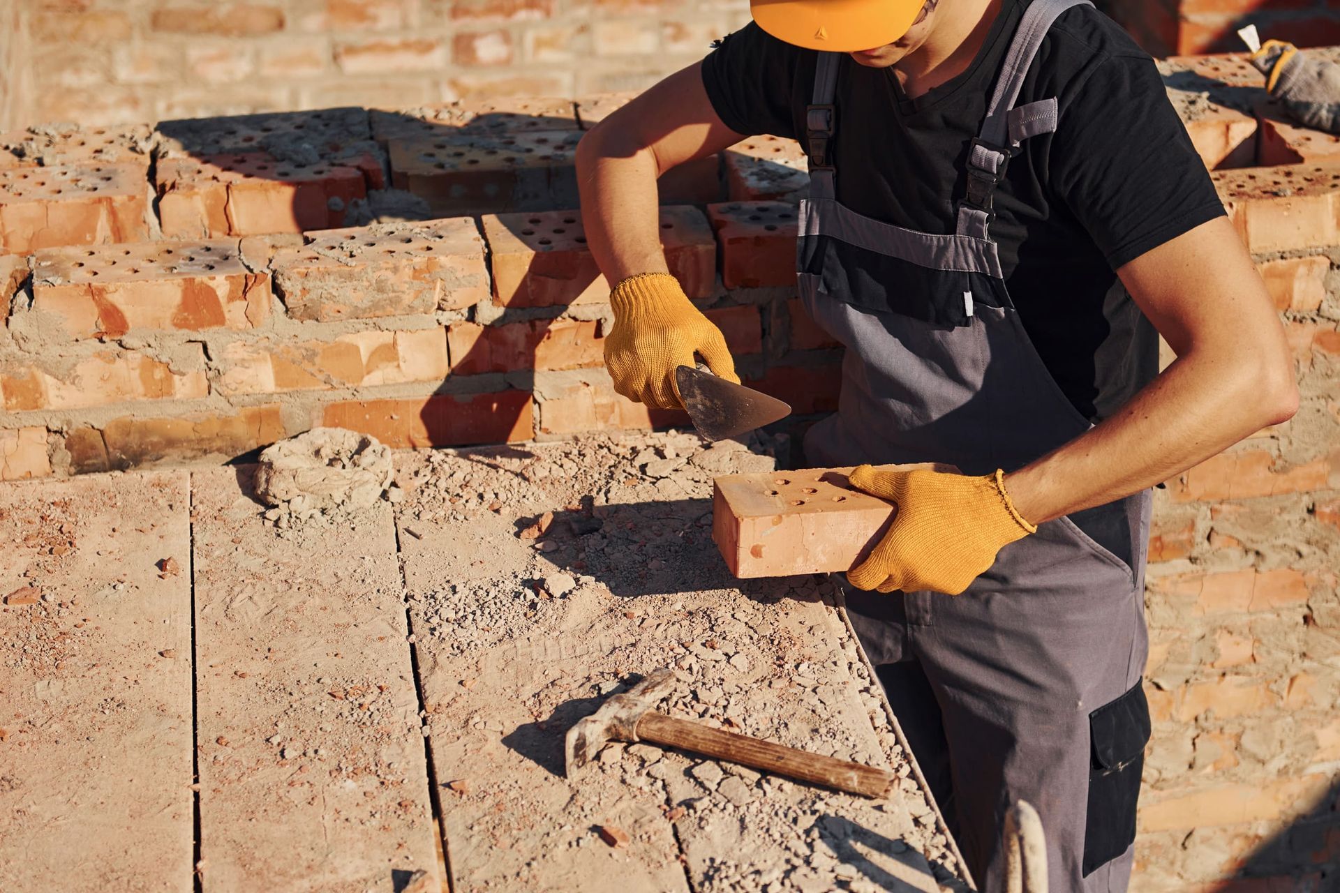 Obrero de la construcción colocando ladrillos con paleta, vistiendo guantes amarillos, overol y casco.