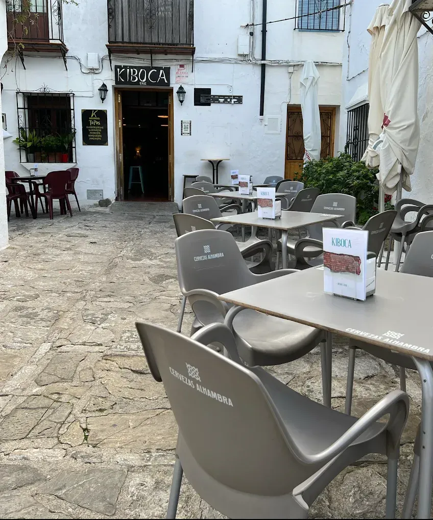 Cafetería al aire libre con mesas y sillas, entrada a un bar etiquetado