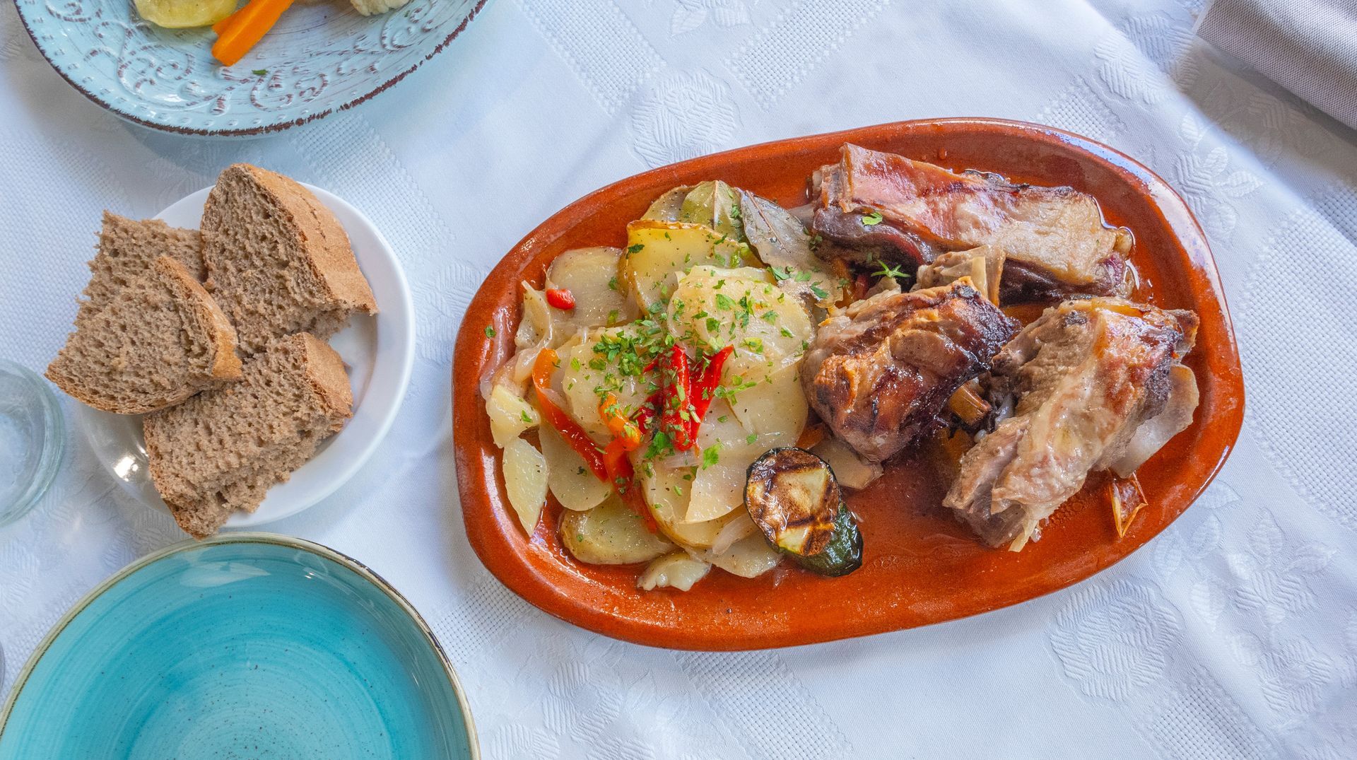 Carne asada con patatas y verduras en plato de barro, servida con pan y guarnición de verduras.