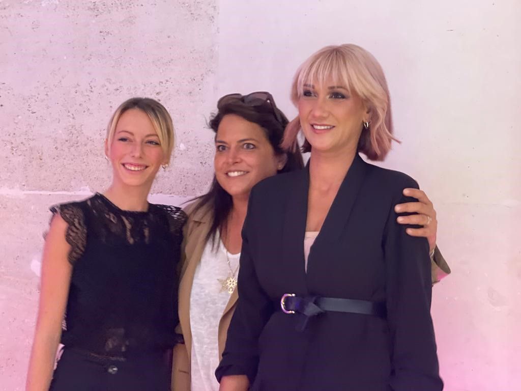 Trois femmes posent ensemble lors d'un événement, souriant devant un mur éclairé en rose.