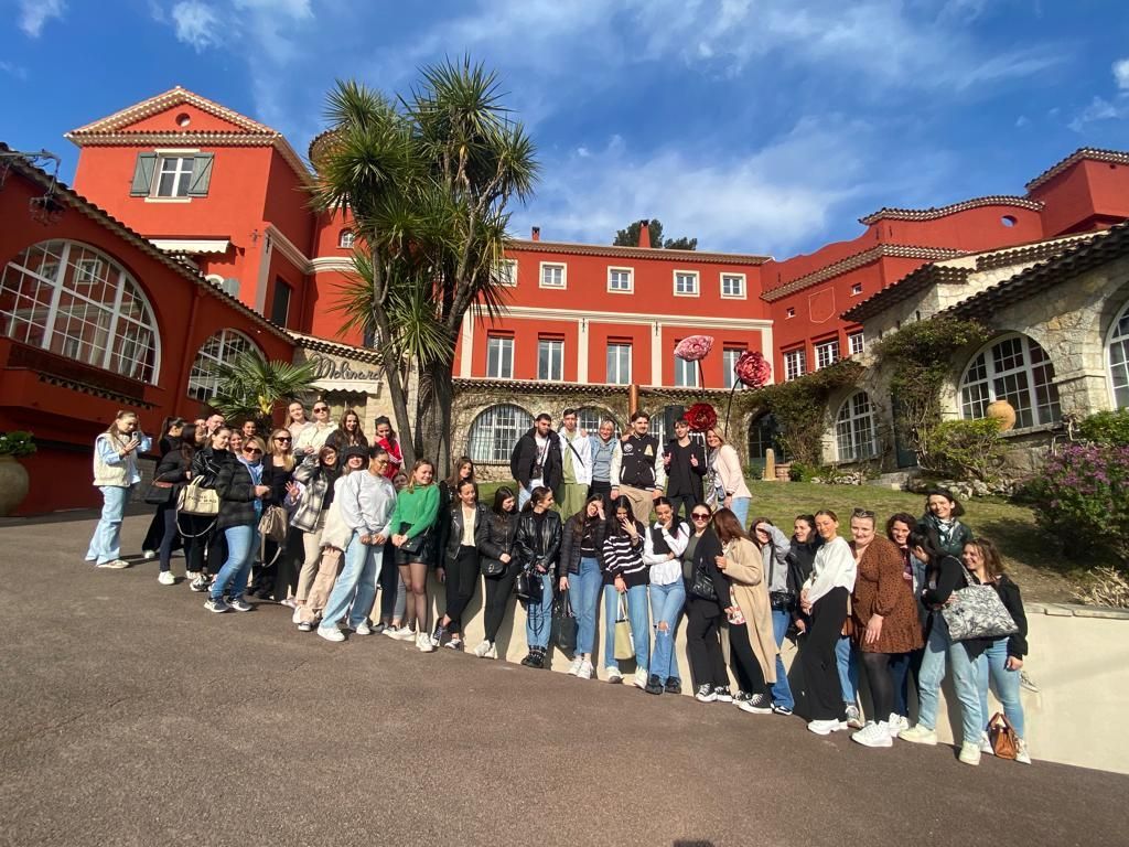 Un groupe de personnes pose devant un bâtiment rouge à arcades, sous un ciel d'un bleu éclatant.