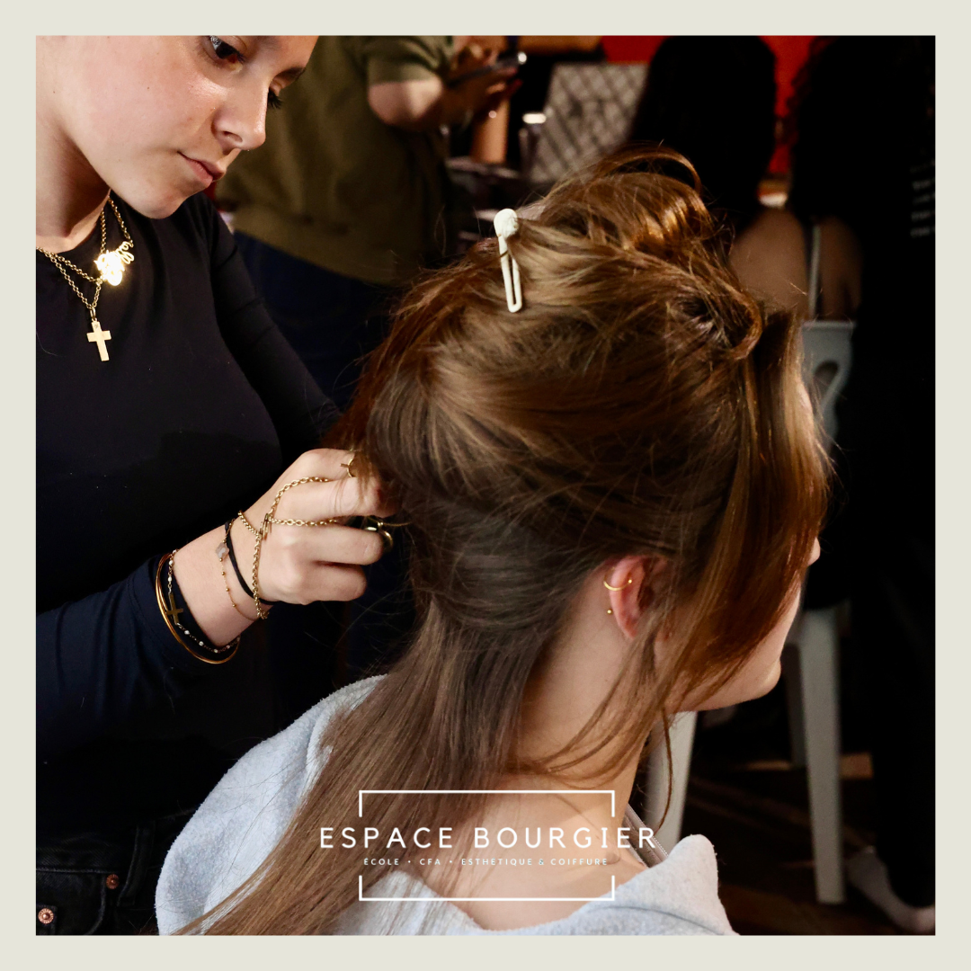 Une coiffeuse réalise une coiffure relevée dans un salon, sa cliente étant assise de profil.