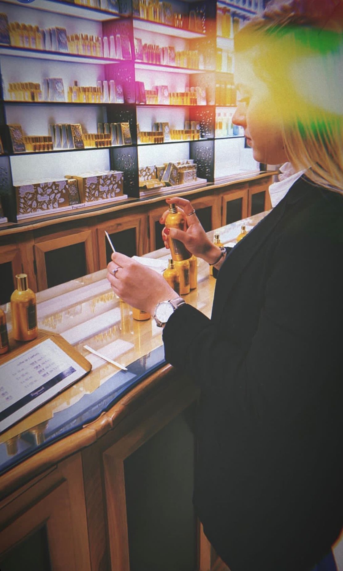 Une personne tenant un flacon de parfumerie, devant des présentoirs de parfums colorés et une lumière vive en arrière-plan.