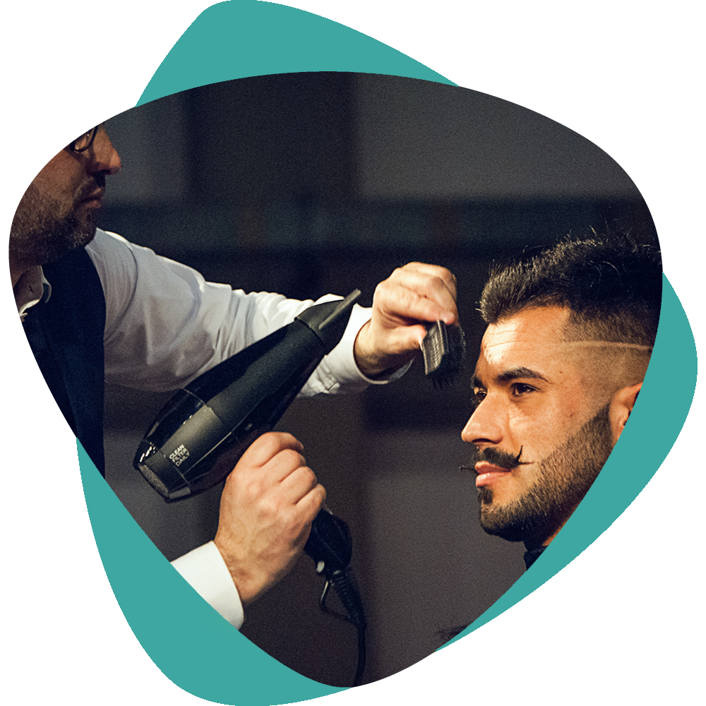 Un coiffeur sèche les cheveux d'un client au sèche-cheveux pendant une coupe, sur fond turquoise.