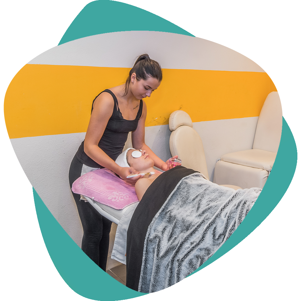Une esthéticienne effectue un massage facial sur une cliente allongée sur une table de soin, devant un décor mural turquoise et jaune.