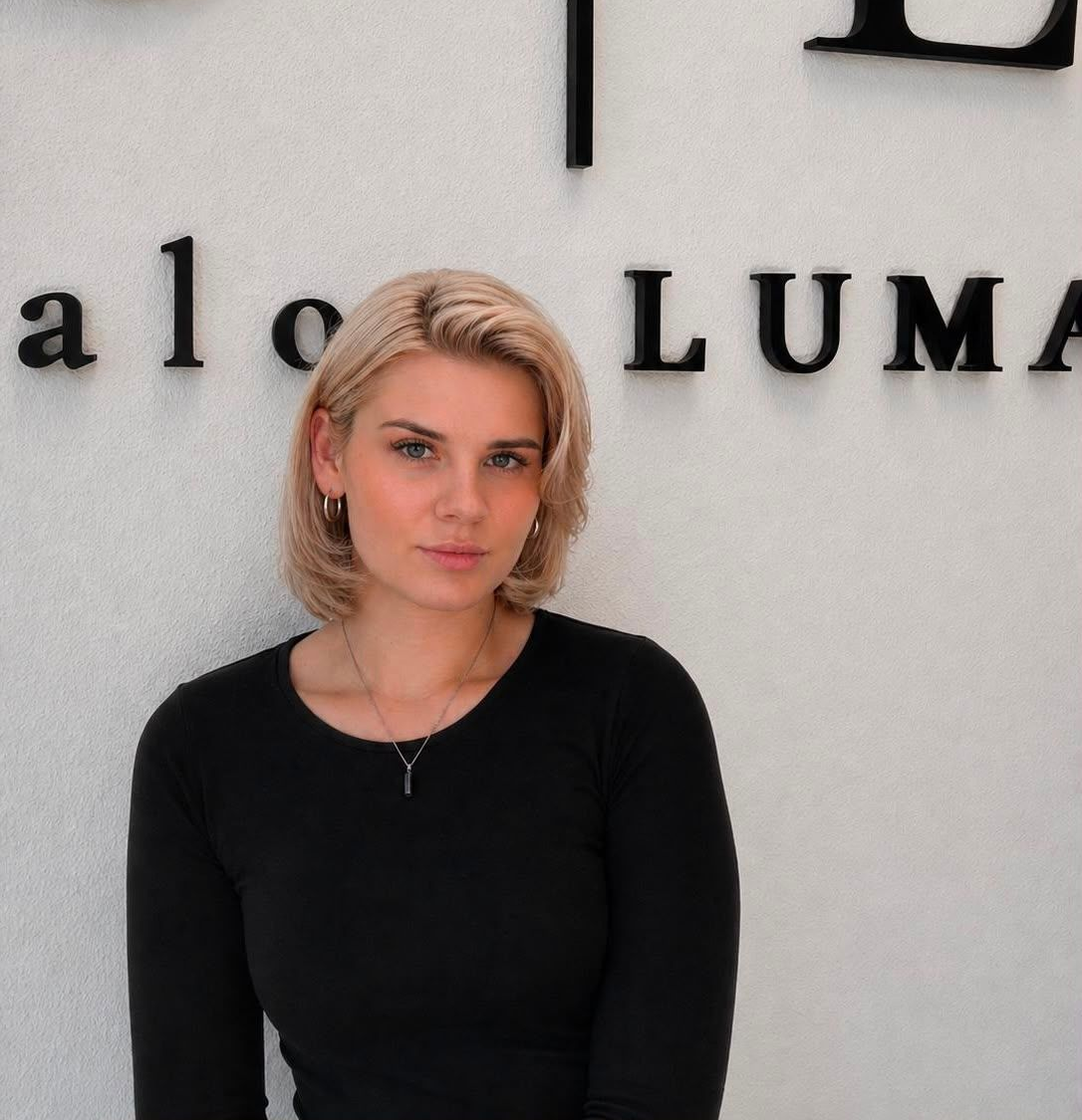 Eine lächelnde Frau sitzt vor dem Schild „S|L Salon LUMA“. Sie trägt ein gemustertes Kleid.