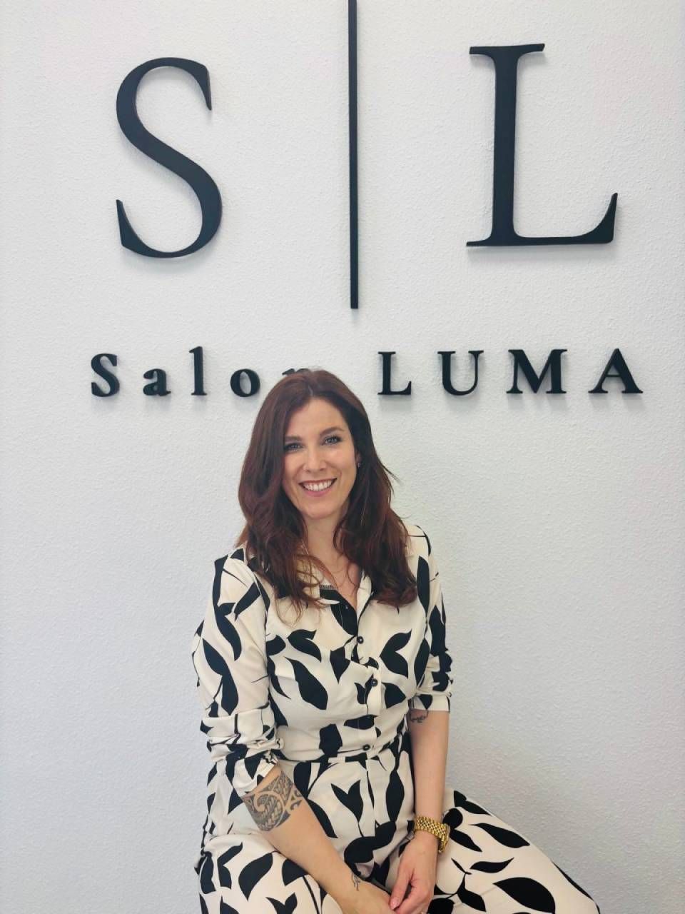 Eine lächelnde Frau sitzt vor dem Schild „S|L Salon LUMA“. Sie trägt ein gemustertes Kleid.