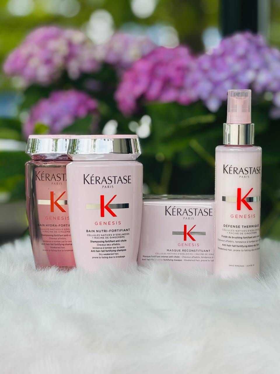 Pinkfarbene Kerastase-Haarpflegeprodukte auf flauschig-weißem Untergrund, im Hintergrund lila und rosafarbene Blumen.