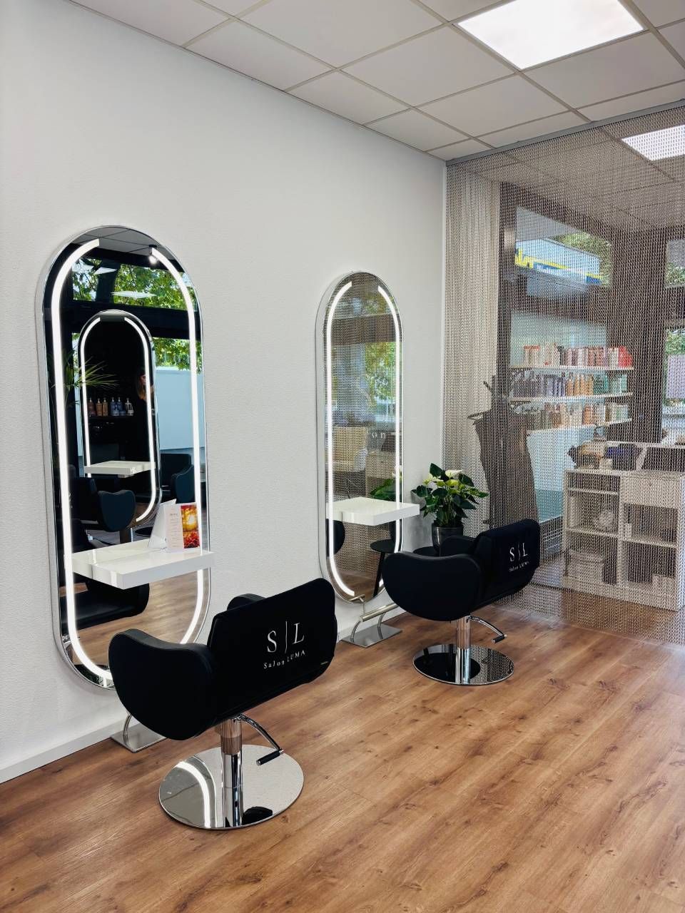 Interieur eines Friseursalons mit zwei schwarzen Stühlen, ovalen Spiegeln mit Beleuchtung und einem hellen Holzboden.
