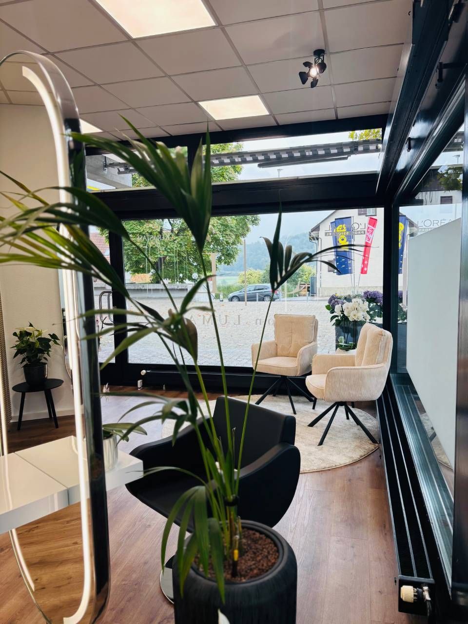 Interieur eines Friseursalons mit zwei cremefarbenen Stühlen, einem schwarzen Stuhl und einer Topfpalme vor einem Fenster.