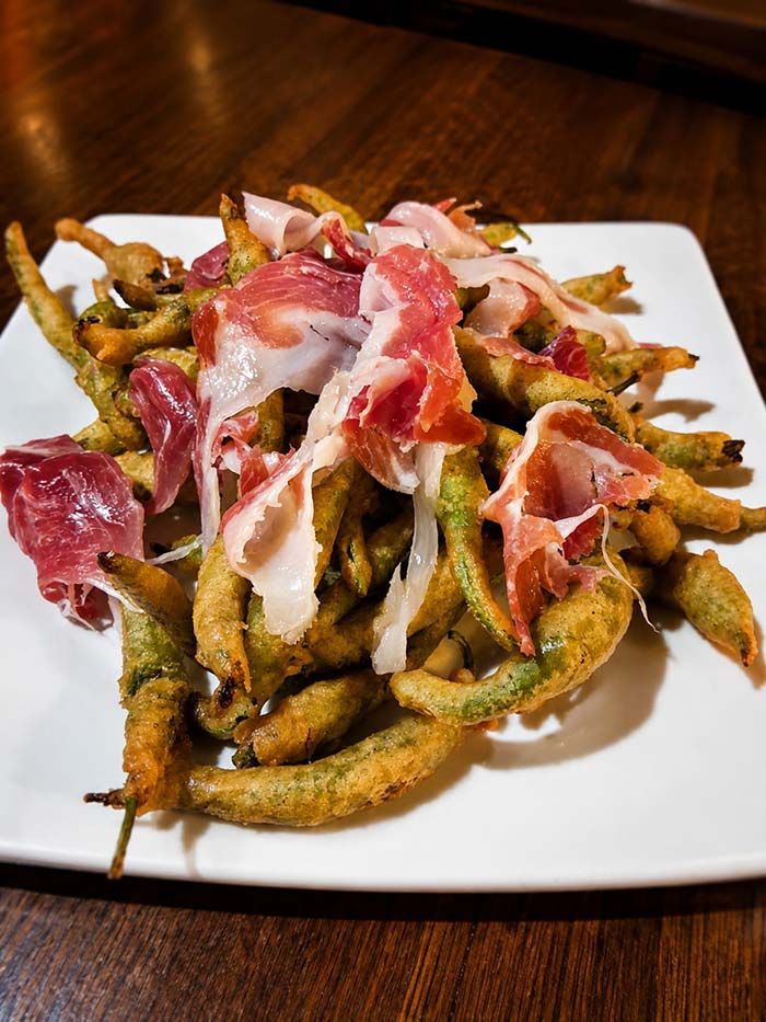Plato de pimientos verdes fritos cubiertos con jamón serrano en lonchas finas sobre un plato blanco.