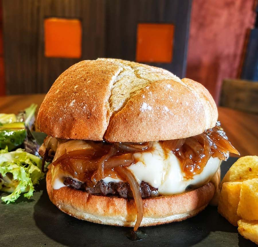 Jugosa hamburguesa con queso y cebolla caramelizada en un panecillo, servida con papas fritas y ensalada en un plato oscuro.