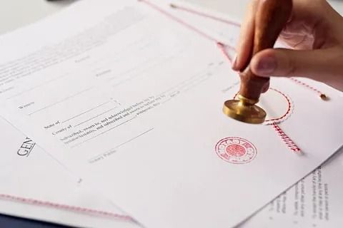 Una mano estampa un sello rojo en documentos oficiales con un hilo rojo, lo que sugiere certificación o aprobación.